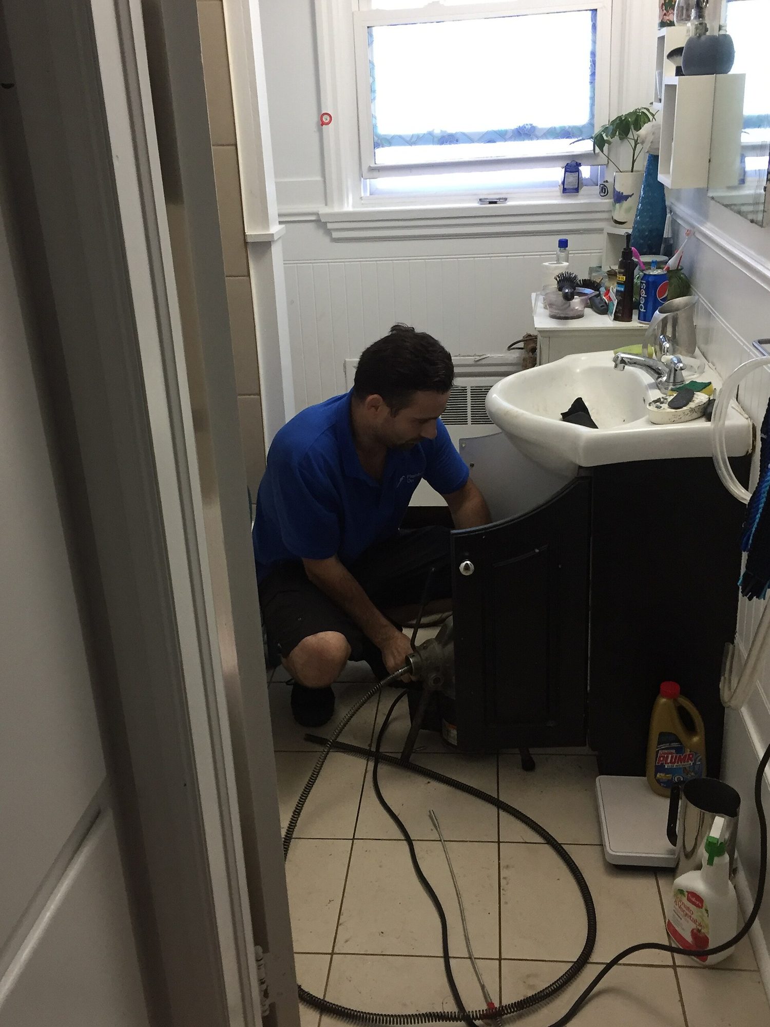 A technician kneeling at a bathroom sink with the drain machine and the whole room in view